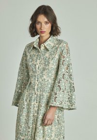 Abito camicia in pizzo verde menta e crema, caratterizzato da un motivo floreale, colletto, chiusura frontale con bottoni e maniche lunghe a campana.