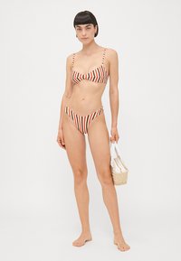 Bikini a righe in rosso e crema, con un top bralette dotato di spalline regolabili e uno slip alta vita. Abbinato a una borsa intrecciata.
