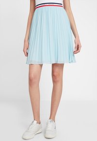 Lichtblauwe plooirok met een elastische tailleband, voorzien van rode en marineblauwe accenten, gemaakt van een doorzichtig materiaal. Draagt met witte sneakers.