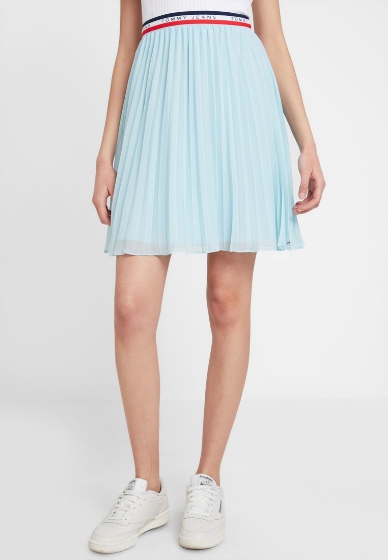 Lichtblauwe plooirok met een elastische tailleband, voorzien van rode en marineblauwe accenten, gemaakt van een doorzichtig materiaal. Draagt met witte sneakers.