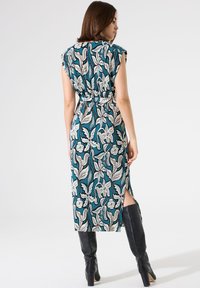 Tealfarbenes Kleid mit floralem Muster, kurzen Ärmeln, tailliertem Bund und seitlichem Schlitz, kombiniert mit kniehohen schwarzen Stiefeln.
