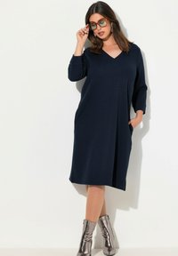 Námořnicky modré šaty s V výstřihem a 3/4 rukávy, které mají boční kapsy a volný střih. Stylizováno s kovovými členkovými botami a oversized brýlemi.