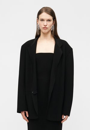 Žena s rovnými vlasmi nosí príliš veľké čierne sako cez čierne šaty bez ramienok a dlhé visiace náušnice z krištáľov na bielom pozadí.