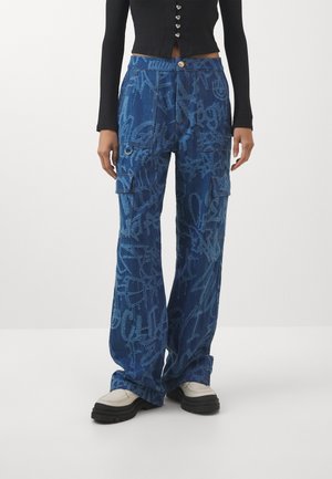 Persona que lleva pantalones cargo vaqueros azules de pierna ancha con estampado estilo graffiti, combinados con zapatos blancos gruesos y una camiseta negra de manga larga.