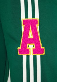 Zelená mikina s velkým, texturovaným růžovým písmenem "A" s žlutým obrysem. Bílé pruhy zdobí strany a vytvářejí sportovní design.