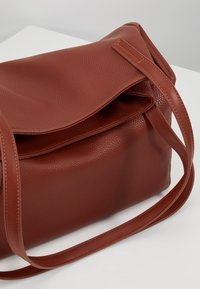 Bruine schoudertas van getextureerd leer met een klepsluiting en een lange riem, voorzien van zichtbare stiksels en een rechthoekige vorm.