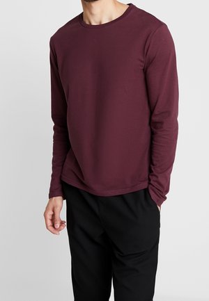 Mann trägt ein einfaches langärmeliges bordeauxrotes Hemd und schwarze Hose, steht mit einer Hand in der Tasche vor einem neutralen Hintergrund.