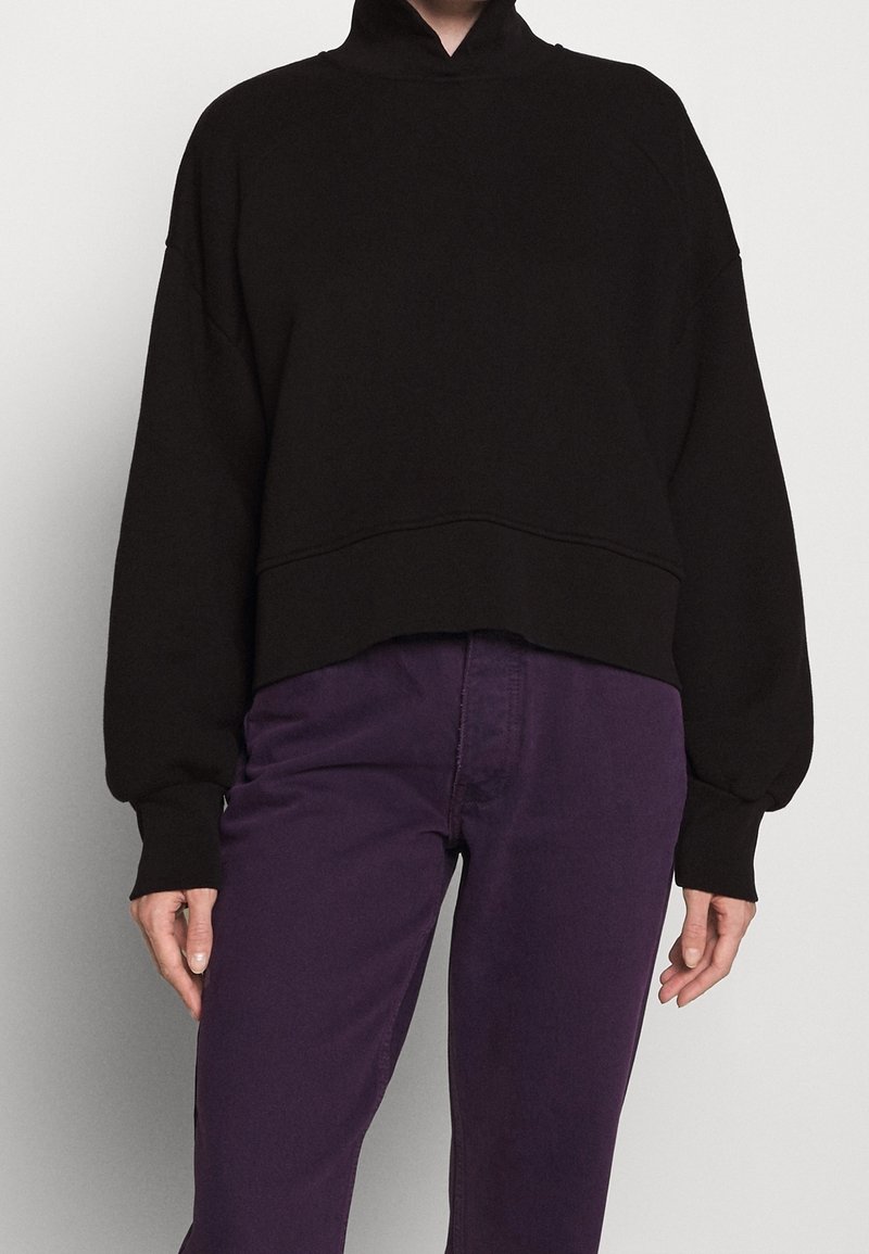Personne portant un sweat court noir à col montant et un pantalon violet taille haute sur un fond clair uni.