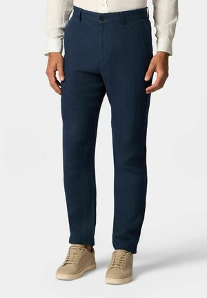 Mężczyzna w dopasowanych granatowych spodniach, beżowych trampkach i białej koszuli na długi rękaw stojący na jednolitym tle.