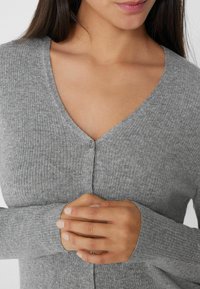 Grauer gerippter Cardigan mit V-Ausschnitt, Knopfverschluss und langen Ärmeln mit taillierten Bündchen. Der Stoff wirkt weich und strukturiert.