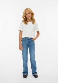 Camiseta blanca de manga corta combinada con unos pantalones vaqueros acampanados de color azul claro, con un solo bolsillo, llevada con zapatos planos negros.