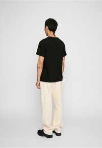 Sort kortærmet t-shirt parret med løstsiddende cremefarvede cargo-bukser og sorte sko, set bagfra mod en ensfarvet baggrund.