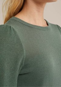 Grön stickad tröja med rund halsringning och puffärmar, med en mjuk textur och subtile detaljer i tyget.