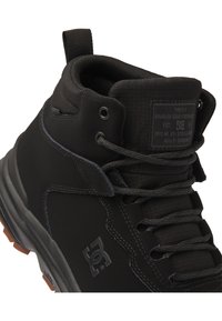 Zapatilla negra de media caña con una parte superior de tela resistente, que presenta cordones negros, un parche de logo y una suela texturizada similar al caucho.