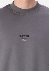 Grå sweatshirt med ribbet runde hals. Har et trykt logo i sort og hvidt på brystet. Blødt, struktureret stof.