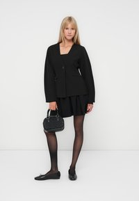 Svart vävd blazer med en enda knapp, kombinerad med en plisserad svart kjol, svarta strumpbyxor och en texturerad svart handväska. Platta svarta skor fulländar looken.