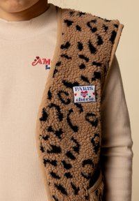 Beige fleece vest met zwarte luipaardvlekken, voorzien van een patch met "PARIS" en "ma chérie." Geribbelde beige top eronder.