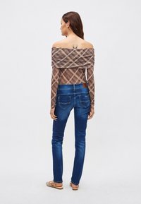 Rudos spalvos languota petnešėlėmis viršutinė dalis su raukštu dizainu ir ilgomis rankovėmis, derinama su mėlynais džinsais, kuriuose yra užpakalinės kišenės ir rudos odos pleistras.