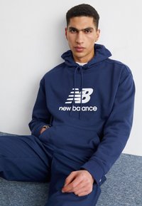 Sötétkék kapucnis pulóver puha anyagból, elöl fehér New Balance logóval. Állítható zsinór, valamint kenguru zsebes részlet.
