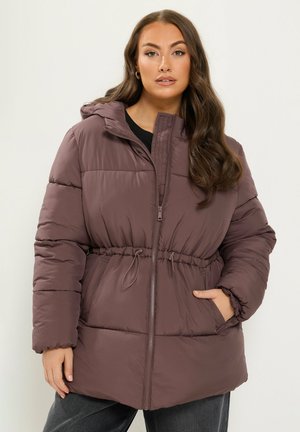 Femme portant une doudoune marron avec une fermeture éclair à l'avant, une capuche, une taille à cordon et les mains dans les poches, debout devant un fond uni.