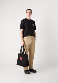 Black canvas tote bag featuring a red label, carried by a model wearing a black t-shirt and beige trousers, with simple black footwear.