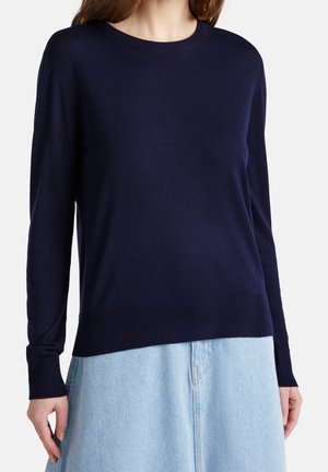 Femme portant un pull à manches longues bleu marine uni, associé à un pantalon en denim bleu clair, debout devant un fond blanc.