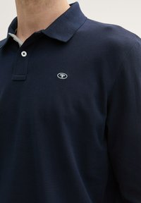 Tumesinine polo-särgid, mis on valmistatud tekstuuritud kangast, kaelusega, kahe nupuga taljes ja väikese logoga rinnal.