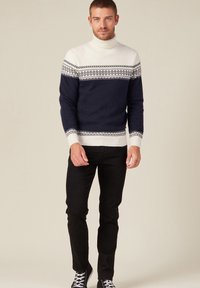 Pull en maille bleu marine foncé avec un col roulé blanc et un motif Fair Isle, présentant des designs géométriques en blanc, gris et accents noirs. Pantalons et chaussures noires.
