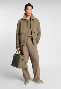 Tan wool blend jacket with large pockets, layered over a green textured knit sweater, paired with beige pleated trousers and a green leather bag.