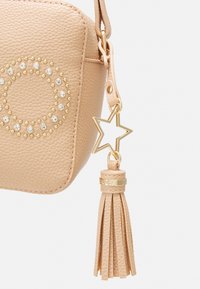 Borsa in finta pelle beige con un design a cerchio con borchie, hardware dorato e un ciondolo a forma di stella con un accento a frangia.