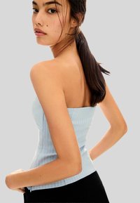 Jeune femme avec de longs cheveux foncés en queue de cheval portant un haut sans manches bleu clair aux rayures noires et un pantalon noir, posant devant un fond uni.