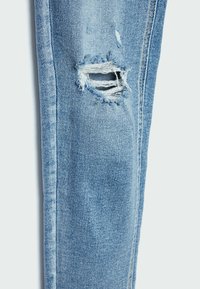 Jeans de mezclilla desgastados de color azul claro con un desgarro prominente y bordes desgastados. Corte estándar con costuras visibles a lo largo de las costuras.