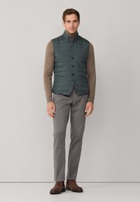 Gesteppte türkisfarbene Weste mit hohem Kragen, Knöpfen vorne und zwei Seitentaschen. Kombiniert mit einem braunen Langarmshirt und grauer Hose.