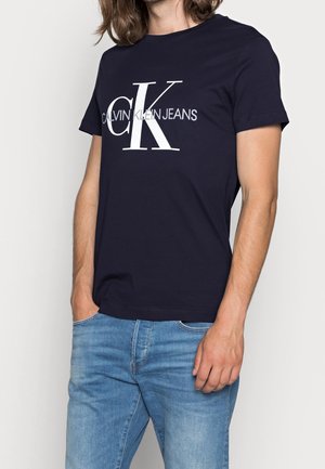 Hombre con camiseta azul marino de Calvin Klein Jeans y vaqueros azul claro, de pie con una mano en el bolsillo contra un fondo blanco.