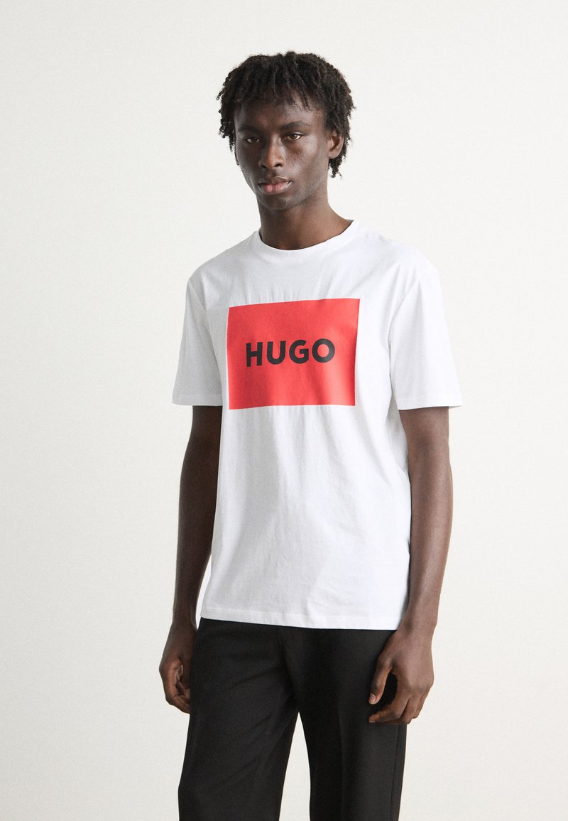 Hvit bomull T-skjorte med en stor rød rektangulær grafikk med teksten "HUGO" i fet svart skrift. Korte ermer og rund hals.