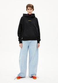 Zwarte hoodie met een voorzak, gele tekst, gecombineerd met lichtblauwe wijde jeans en rode sneakers. Glad materiaal, comfortabele pasvorm.