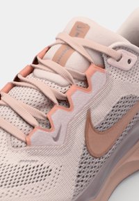 Zapatilla deportiva de color rosa claro con parte superior de malla, acentos angulares en un rosa más oscuro y un logotipo prominente en el lateral. Presenta cordones texturizados.