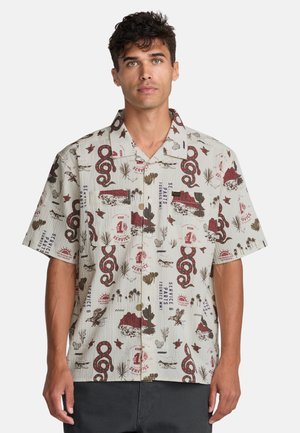 Mężczyzna ubrany w beżową koszulę z krótkim rękawem, zapinaną na guziki, z czerwonymi i brązowymi motywami pustynnymi, w tym węże, kaktusy i wzory z tekstem.