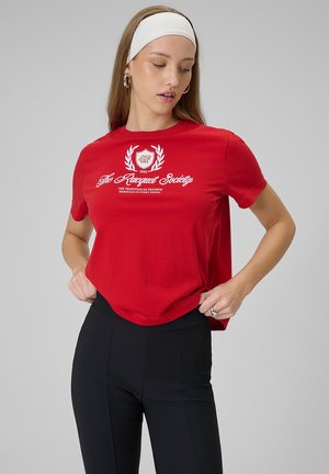 Młoda kobieta poprawia czerwoną koszulkę z białym napisem „The Racquet Society” i emblematem, ubrana w czarne spodnie i białą opaskę na głowę na szarym tle.