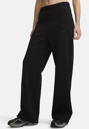 Person wearing loose black Under Armour pants with zippered side pockets and black athletic shoes.