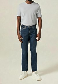 Homme portant un t-shirt blanc uni, un jean droit bleu foncé et des baskets blanches, debout devant un fond neutre.