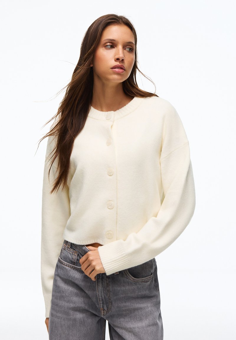 Femme aux longs cheveux bruns portant un cardigan crème boutonné et un jean gris taille haute, debout devant un fond blanc.