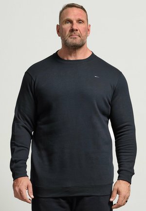 Skægget mand med kort hår iført en ensfarvet mørk trøje med lange ærmer, står mod en lys ensfarvet baggrund og kigger fremad.