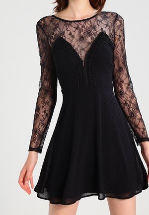 Robe noire avec manches longues en dentelle transparente et haut de poitrine, taille cintrée, jupe évasée et bordure détaillée sur le devant du corsage en forme de V.