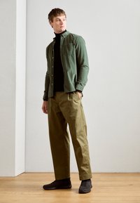 Camisa verde de botones sobre un cuello de tortuga negro, combinada con pantalones en un tono oliva claro y zapatos oscuros. Texturas suaves, corte relajado. Fondo de suelo de madera.