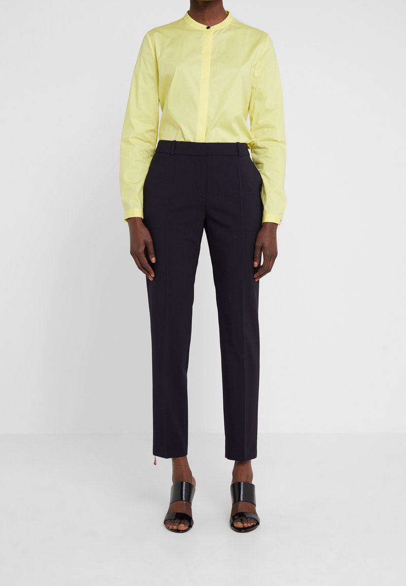 Personne portant une chemise jaune à manches longues, un pantalon noir cintré et des sandales noires à bout ouvert sur un fond blanc uni.