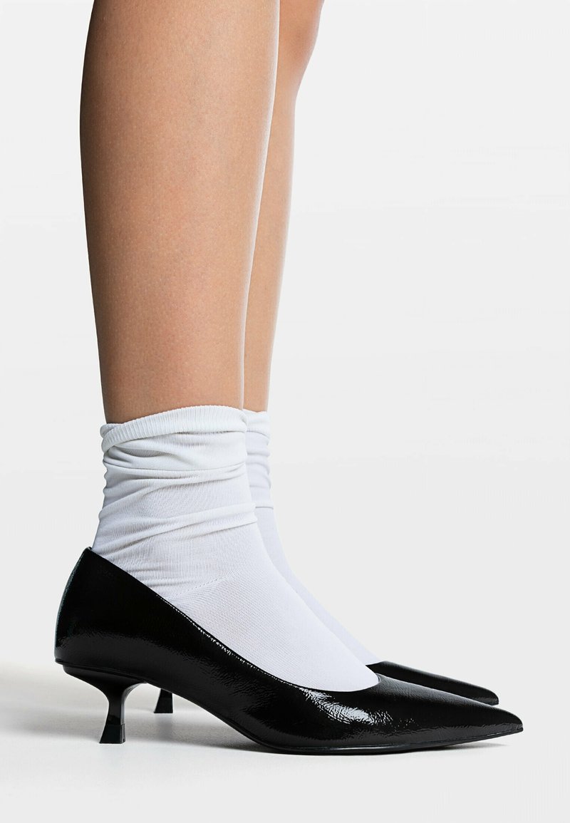Escarpins en cuir verni noir avec un bout pointu et un petit talon, portés avec des chaussettes blanches à la cheville. Texture lisse et finition brillante.