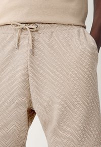 Bézs mintás melegítőnadrágok texturált geometriai mintával, elasztikus derékrésszel, húzózsinórral és fémes végződéssel a húzózsinóron.