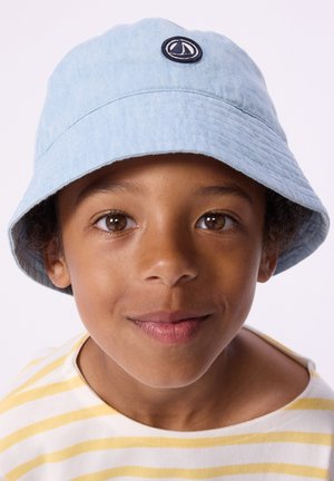 Chapeau bob bleu clair en tissu texturé, avec un logo circulaire bleu marine à l'avant. Le modèle porte une chemise à rayures jaunes en dessous.