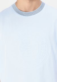 Tricou albastru deschis cu guler ribatat alb și negru, având un logo Lacoste reliefat și un design de frunză pe față.
